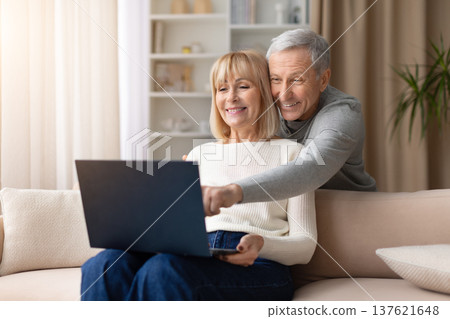 Happy senior couple sitting on sofa together using laptop at home Happy senior couple sitting on sofa together using laptop at home 137621648