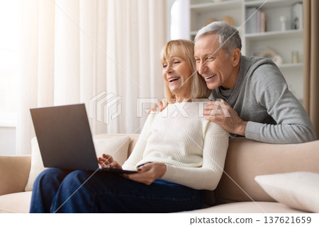 Senior couple sitting together on sofa at home using laptop and smiling Senior couple sitting together on sofa at home using laptop and smiling 137621659