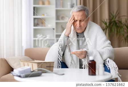 Senior man sitting on sofa at home checking thermometer while feeling unwell Senior man sitting on sofa at home checking thermometer while feeling unwell 137621662