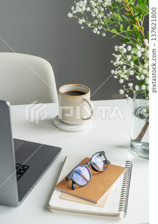A coffee mug, laptop, notepad, and glasses on a white table with gypsophila flowers in a glass vase A coffee mug, laptop, notepad, and glasses on a white table with gypsophila flowers in a glass vase 137621800