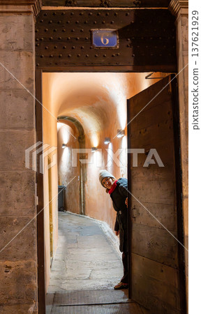 Lyon, france. December 29, 2023. Woman peeking from an open doorway, revealing a long arched traboule passage, an architectural feature in lyon, france Lyon, france. December 29, 2023. Woman peeking from an open doorway, revealing a long arched traboule passage, an architectural feature in lyon, france 137621929