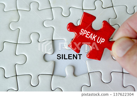 Hand positioning a red speak up puzzle piece above the word silent on a white jigsaw puzzle background Hand positioning a red speak up puzzle piece above the word silent on a white jigsaw puzzle background 137622304