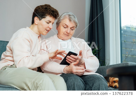 Adult woman explaining her senior mother how to use smartphone. Education for aged. Retired lady discovering mobile technology for calling, texting, buying online, playing games, using medical apps Adult woman explaining her senior mother how to use smartphone. Education for aged. Retired lady discovering mobile technology for calling, texting, buying online, playing games, using medical apps 137622378