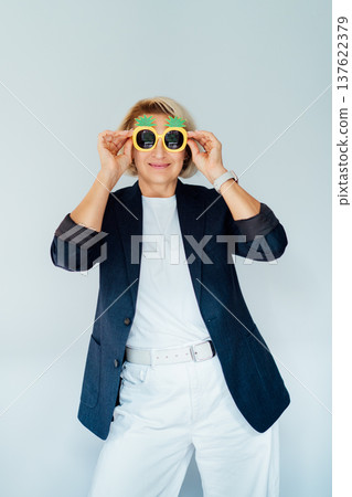 Middle age woman in blazer and white outfit posing in pineapple sunglasses over isolated white background, smiling to the camera. Relaxed business woman. Vertical card. 137622379