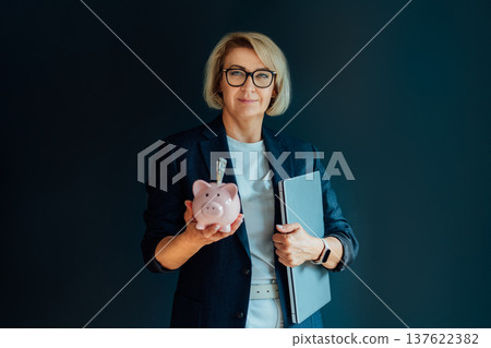 Stylish 50's confident mature businesswoman holding piggy bank with money and laptop in other hand on dark background. Financial advisor, accounting, tax, investment, pension specialist. Stylish 50's confident mature businesswoman holding piggy bank with money and laptop in other hand on dark background. Financial advisor, accounting, tax, investment, pension specialist. 137622382