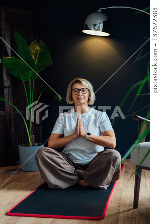 Middle aged woman practicing yoga, meditation at home, sitting in Namaste position on floor mat in modern Scandi dark interior home. Meditating, relaxed, eyes closed. Mental health and well being. Middle aged woman practicing yoga, meditation at home, sitting in Namaste position on floor mat in modern Scandi dark interior home. Meditating, relaxed, eyes closed. Mental health and well being. 137622383