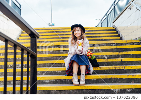 Portrait of young smiling woman with fresh tulips and coffee cup sitting on stairs at the city street enjoying the moment. Urban street fashion. Springtime yellow mood. Happiness and positive vibes Portrait of young smiling woman with fresh tulips and coffee cup sitting on stairs at the city street enjoying the moment. Urban street fashion. Springtime yellow mood. Happiness and positive vibes 137622386
