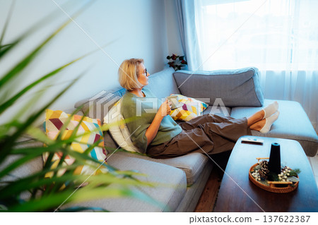 Middle aged woman relaxing with cup of hot drink on sofa in hygge interior home. Cozy comfortable winter or autumn weekends. Take a break and relax. Lady enjoy calm mood, well being alone Middle aged woman relaxing with cup of hot drink on sofa in hygge interior home. Cozy comfortable winter or autumn weekends. Take a break and relax. Lady enjoy calm mood, well being alone 137622387