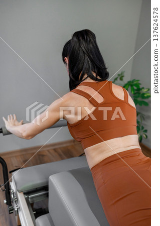 Woman stretching arms on pilates reformer wearing burnt orange activewear in bright studio. Pilates reformer, stretching, studio, orange, athletic. Woman stretching arms on pilates reformer wearing burnt orange activewear in bright studio. Pilates reformer, stretching, studio, orange, athletic. 137624578
