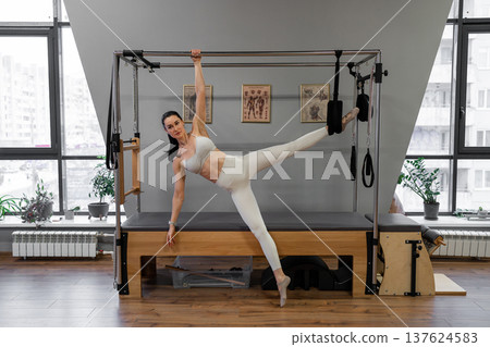 Active woman doing side leg raise on pilates reformer near window, with sunny light in studio. Side leg raise, pilates reformer, active, sunny, exercise. Active woman doing side leg raise on pilates reformer near window, with sunny light in studio. Side leg raise, pilates reformer, active, sunny, exercise. 137624583