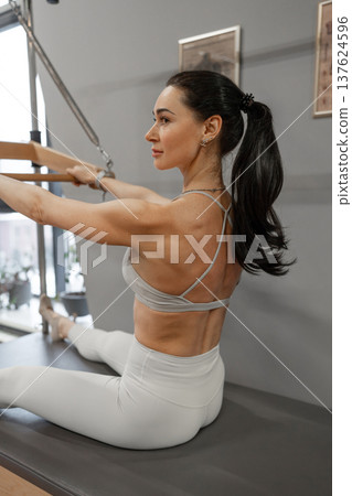 Focused young woman stretching arms with resistance bars on pilates reformer machine. Stretching arms, resistance bars, focus, pilates, fitness. Focused young woman stretching arms with resistance bars on pilates reformer machine. Stretching arms, resistance bars, focus, pilates, fitness. 137624596