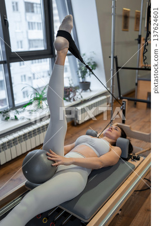 Woman in sportswear holding pilates pose on reformer machine with legs raised in bright studio. Pilates, legs raised, sportswear, reformer, stretching. Woman in sportswear holding pilates pose on reformer machine with legs raised in bright studio. Pilates, legs raised, sportswear, reformer, stretching. 137624601