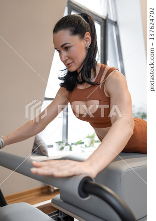 Athletic woman in orange sportswear doing swan and working out on pilates reformer in bright indoor studio. Pilates workout, studio, orange sportswear, athletic, training. Athletic woman in orange sportswear doing swan and working out on pilates reformer in bright indoor studio. Pilates workout, studio, orange sportswear, athletic, training. 137624702
