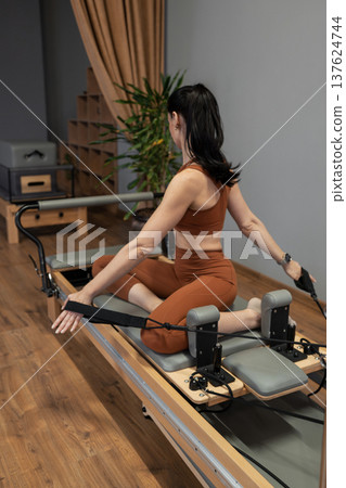 Woman stretching with long resistance strap during pilates exercise routine in bright gym. Resistance strap, stretching, pilates, routine, gym. Woman stretching with long resistance strap during pilates exercise routine in bright gym. Resistance strap, stretching, pilates, routine, gym. 137624744