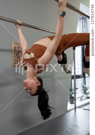 Woman lying back, arching and hanging from pilates bar in gymnasium during advanced workout. Arching, hanging, pilates bar, gymnasium, advanced workout. 137624746