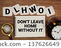 DLHWOI Don't Leave Home Without It text on notepad with magnifying glass and alarm clock on wooden background 137626649
