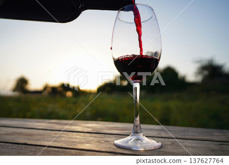 Red wine is poured from a bottle into a glass. The process of decanting wine outdoors at sunset. Red wine is poured from a bottle into a glass. The process of decanting wine outdoors at sunset. 137627764
