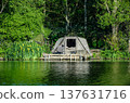 Fishing tent camp located directly at the edge of calm lake water surrounded by green vegetation and reeds. A quiet outdoor environment showing a peaceful lakeside fishing lifestyle scene. 137631716