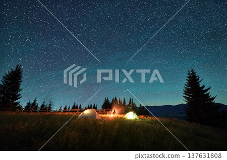 Silhouette of couple hikers resting by campfire under star-studded sky, near illuminated tents at campsite. Night camping in mountains, night sky create magical and peaceful atmosphere. Silhouette of couple hikers resting by campfire under star-studded sky, near illuminated tents at campsite. Night camping in mountains, night sky create magical and peaceful atmosphere. 137631808