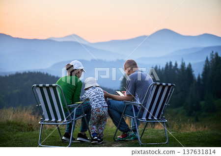Family of three sits on foldable chairs, enjoying peaceful sunset in mountains, share time together in nature. Child stands between parents. Layered mountain ranges and soft, colorful sky. 137631814