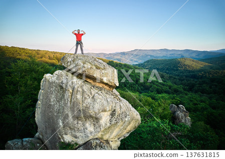 Aerial view of climber standing atop rock formation at Dovbush Rocks in Carpathian mountains, Ukraine. Sun sets, casting warm glow over lush, green forest, distant hills, creating breathtaking view. 137631815