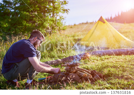 Man tending to campfire on grassy field with yellow tent and backpack nearby. Smoke rises from fire as he works, surrounded by lush greenery and distant tree line under clear blue sky. Man tending to campfire on grassy field with yellow tent and backpack nearby. Smoke rises from fire as he works, surrounded by lush greenery and distant tree line under clear blue sky. 137631816