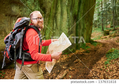 Happy bearded tourist man studies map while stands in front of large rock in dense forest. Smiling traveler with grey backpack and glasses, planning outdoor adventure, hiking or climbing route. Happy bearded tourist man studies map while stands in front of large rock in dense forest. Smiling traveler with grey backpack and glasses, planning outdoor adventure, hiking or climbing route. 137631818