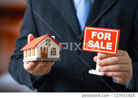 A residential building and a For Sale sign held by a man in a suit. Real estate for sale and property marketing. Real estate agent promoting housing investment and home ownership opportunities. A residential building and a For Sale sign held by a man in a suit. Real estate for sale and property marketing. Real estate agent promoting housing investment and home ownership opportunities. 137631928