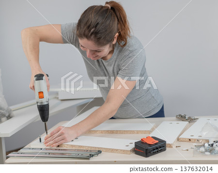 A Caucasian woman uses a screwdriver to tighten a screw while assembling a cabinet. A Caucasian woman uses a screwdriver to tighten a screw while assembling a cabinet. 137632014
