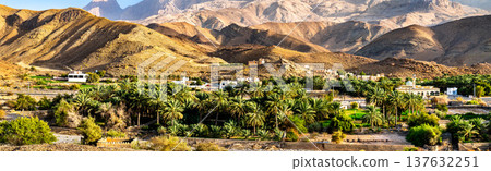 A wide panoramic view of the lush palm oasis and buildings in Al Ayn village and Al Hajar Mountains in Oman A wide panoramic view of the lush palm oasis and buildings in Al Ayn village and Al Hajar Mountains in Oman 137632251