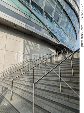 Golden afternoon light streams across the granite steps and steel railings, drawing the eye upward. This grand entrance leads to a massive concrete anchor, crowned by a dramatic glass facade that 137632276