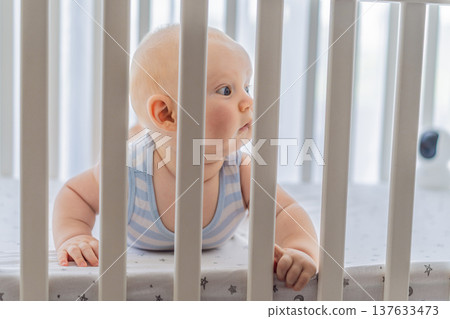 Sad baby looking through the bars of a crib, expressing longing and curiosity. Emotional scene symbolizing the need for freedom and exploration in early childhood. Infant development, autonomy and Sad baby looking through the bars of a crib, expressing longing and curiosity. Emotional scene symbolizing the need for freedom and exploration in early childhood. Infant development, autonomy and 137633473