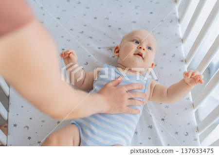 Mother sitting beside the crib gently stroking her baby lying in a cozy bed at home. Warm intimate moment filled with love, trust and calm. Motherhood, infant comfort and emotional bonding concept 137633475