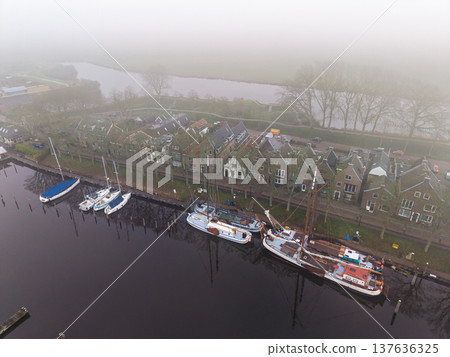 Atmospheric aerial view of traditional sailboats and yachts moored along a canal in a historic Dutch town. Thick morning fog creates a moody, serene European heritage scene. 137636325