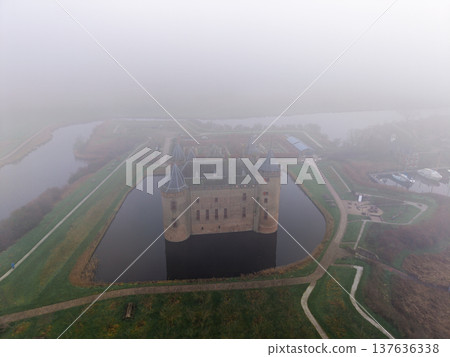 Aerial shot of medieval Muiderslot Castle shrouded in thick fog. Historic brick fortress with towers surrounded by a water moat and misty gardens in the Netherlands. Moody morning atmosphere. Aerial shot of medieval Muiderslot Castle shrouded in thick fog. Historic brick fortress with towers surrounded by a water moat and misty gardens in the Netherlands. Moody morning atmosphere. 137636338