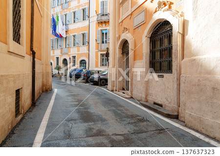 Narrow street with buildings in Rome during daytime with parked cars and flags 137637323