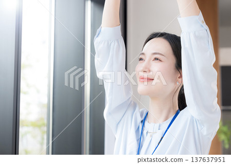 A businesswoman takes a break by the window of her office, stretching and relaxing amidst the fresh greenery of spring. She looks refreshed and invigorated. 137637951