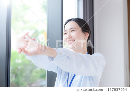 A businesswoman takes a break by the window of her office, stretching and relaxing amidst the fresh greenery of spring. She looks refreshed and invigorated. A businesswoman takes a break by the window of her office, stretching and relaxing amidst the fresh greenery of spring. She looks refreshed and invigorated. 137637954
