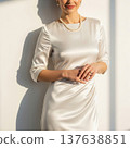 Elegant Woman in Cream Satin Dress Posing in Sunlight Against White Wall 137638851