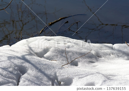 Melting snowdrifts at the riverbank in spring, showcasing the transition from winter to a new season with water reflections and scattered branches Melting snowdrifts at the riverbank in spring, showcasing the transition from winter to a new season with water reflections and scattered branches 137639155