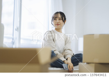 A young woman sitting on a bed mattress surrounded by cardboard boxes. Moving image. 137643411