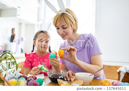 A mother and daughter wearing bunny ears are happily decorating Easter eggs together in a bright kitchen setting, surrounded by colorful eggs and craft supplies A mother and daughter wearing bunny ears are happily decorating Easter eggs together in a bright kitchen setting, surrounded by colorful eggs and craft supplies 137645011