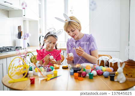 A mother and daughter wearing bunny ears are happily decorating Easter eggs with wax and brushes in a bright kitchen setting A mother and daughter wearing bunny ears are happily decorating Easter eggs with wax and brushes in a bright kitchen setting 137645025