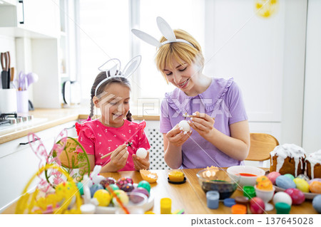 A mother and daughter wearing bunny ears are happily decorating Easter eggs together at a kitchen table with colorful paints and brushes 137645028
