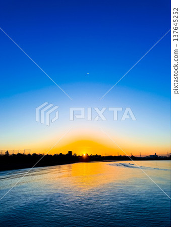A scene of a sunset sinking into the sea and a vast blue sky, with orange light reflecting on the water's surface. 137645252