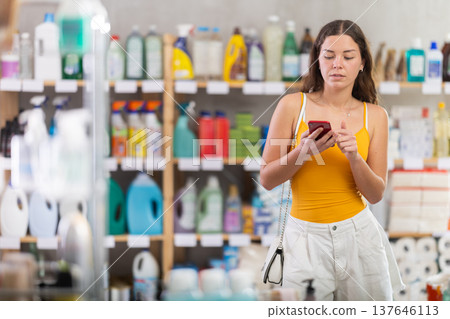 Female shopper searches for necessary goods on the shelves of a supermarket, checking list on her smartphone Female shopper searches for necessary goods on the shelves of a supermarket, checking list on her smartphone 137646113