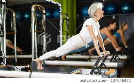 Senior woman standing in plank on pilates reformer, perform exercises Senior woman standing in plank on pilates reformer, perform exercises 137646263