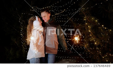 Romantic couple sharing festive moment, holding sparklers near illuminated pine tree, creating intimate new year's eve celebration under twinkling string lights 137646489