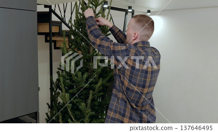 Man decorating contemporary christmas tree, positioning ornaments and branches with precision, creating festive home interior ambiance Man decorating contemporary christmas tree, positioning ornaments and branches with precision, creating festive home interior ambiance 137646495
