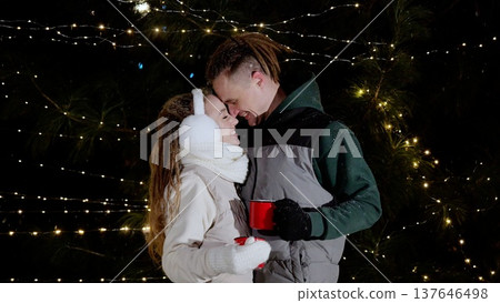 Happy young couple smiling and touching foreheads in front of christmas tree lights, holding red mugs, wearing winter clothes, enjoying christmas time together 137646498